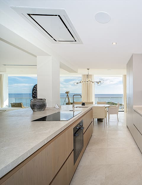 Galley kitchen with light grey handleless cabinets, stone countertops, and built-in appliances, opening to a dining area overlooking the sea.