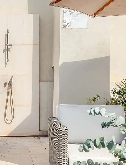 An outdoor shower fixture mounted on a light stone wall on a sunny terrace with a large potted agave plant.