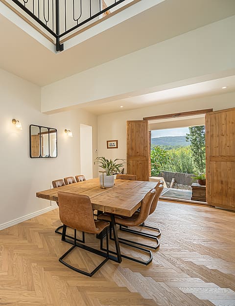 Dining room featuring a rustic wooden table, leather chairs, herringbone wood floors, and open doors leading to a garden.
