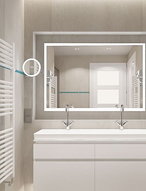 A modern bathroom featuring a white double vanity, large rectangular mirror with integrated lighting, and white heated towel rails against neutral stone-tiled walls.