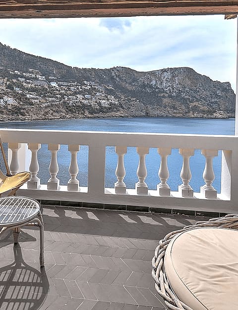 Covered terrace with stone floor, two yellow folding chairs, and a rattan lounge seat overlooking the ocean and mountains.
