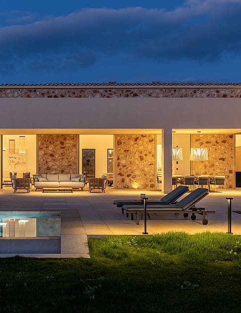 Exterior view of a stone finca with a pool and terrace illuminated at twilight, featuring outdoor furniture and lawn.