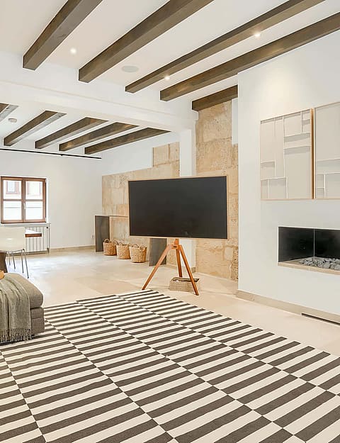 Wide angle view of an open-plan living room featuring exposed dark wood ceiling beams, white walls, a stone-surround fireplace, and a large patterned area rug.