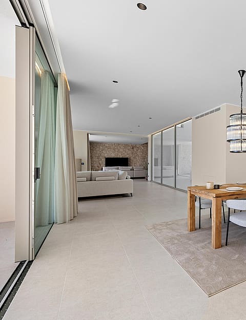 Open-plan living and dining area featuring floor-to-ceiling windows, a wooden dining table, fabric chairs, and designer lighting.