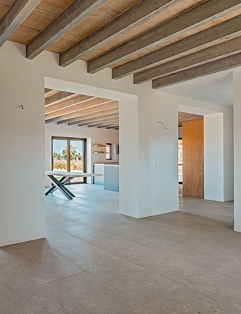 Empty open-plan living room in a finca with white walls, exposed wood ceiling beams, and stone tile floors.