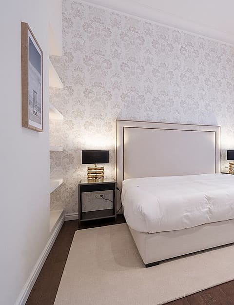 A bright bedroom featuring a queen bed, beige rug, white patterned wallpaper, and sheer curtains over a window.
