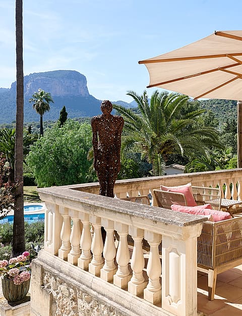 A stone terrace with a beige umbrella and outdoor seating, overlooking the Alaro mountains under clear daylight.