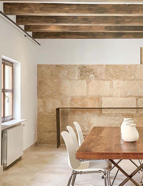 Dining area with a long wooden table, modern white chairs, exposed timber ceiling beams, and textured stone block wall.