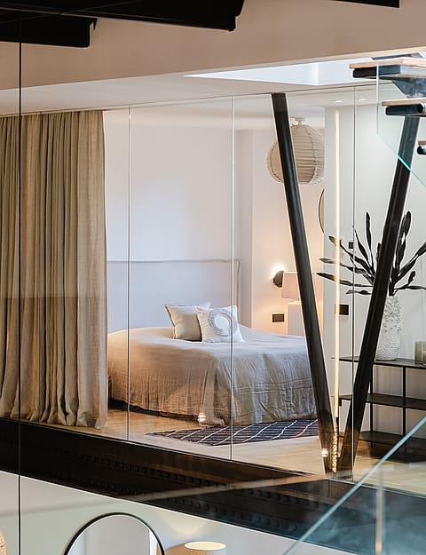 Interior view showing a master bedroom partially enclosed by glass partitions, featuring a modern floating staircase with glass railings and dark steel beams.