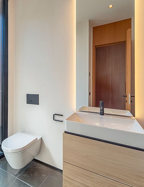 A modern guest bathroom featuring a wall-mounted toilet, wooden vanity with a white sink, backlit mirror, and floor-to-ceiling glass panel looking out to a garden.