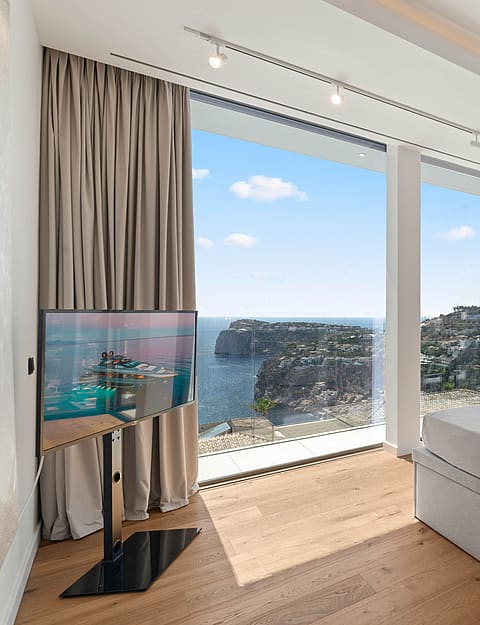 Modern bedroom featuring a king bed facing floor-to-ceiling windows overlooking a coastal cliff and sea, with light oak floors.