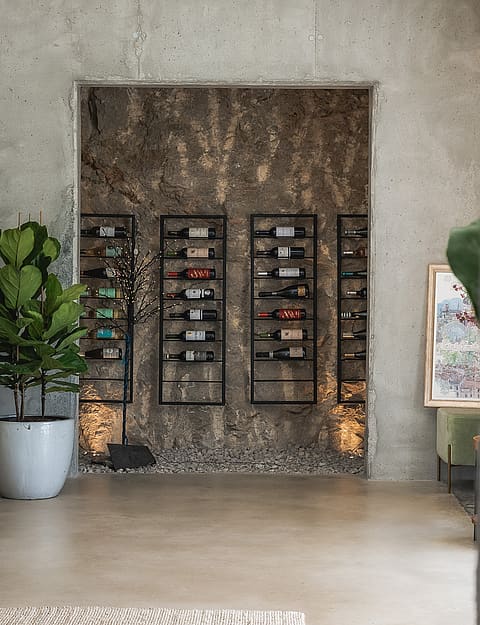 Minimalist wine storage area featuring black metal racks against an exposed rock wall and raw concrete finishes.