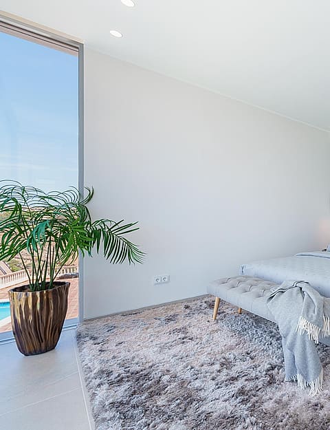 Modern bedroom with grey upholstered bed, glass-paneled en-suite bathroom, and large window overlooking a terrace.