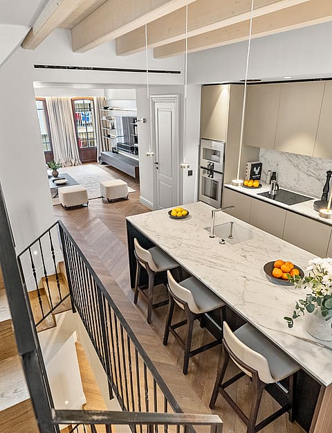 High-angle view of a modern kitchen featuring a large marble island, chevron oak floors, and stairs with metal railings.