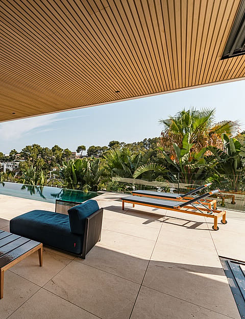 A covered terrace with a wood-slatted ceiling, limestone flooring, and blue outdoor lounge furniture overlooking a swimming pool.