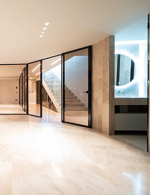 An indoor swimming pool with blue water and beige travertine flooring, adjacent to a glass-enclosed staircase.