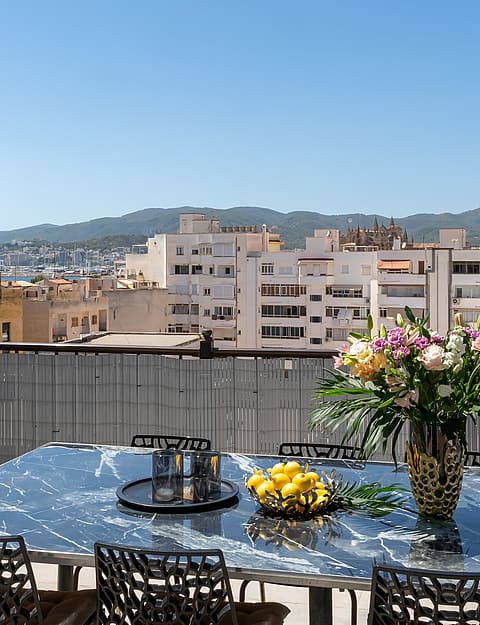 A rooftop terrace with a reflective glass dining table, black wicker chairs, and a flower vase, overlooking Old Town buildings.