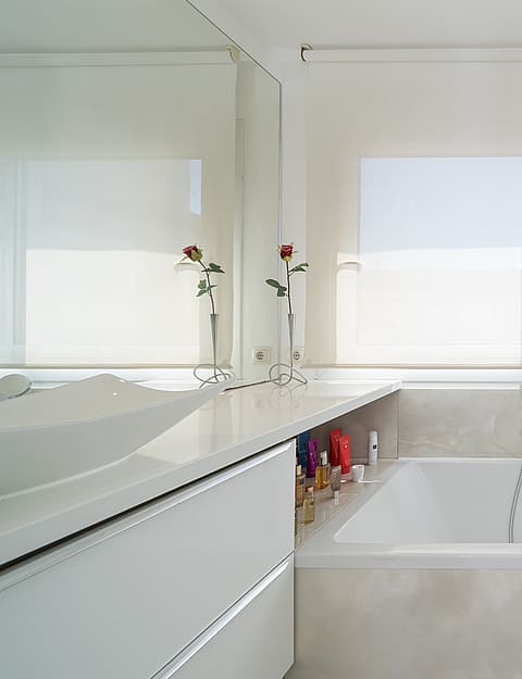 A white bathroom featuring a deep rectangular soaking tub, a wide vanity, and a large window with a roller blind.
