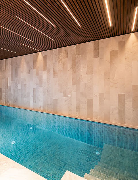 Indoor pool with turquoise water, neutral stone wall cladding, and dark wood slat ceiling with recessed linear lighting.