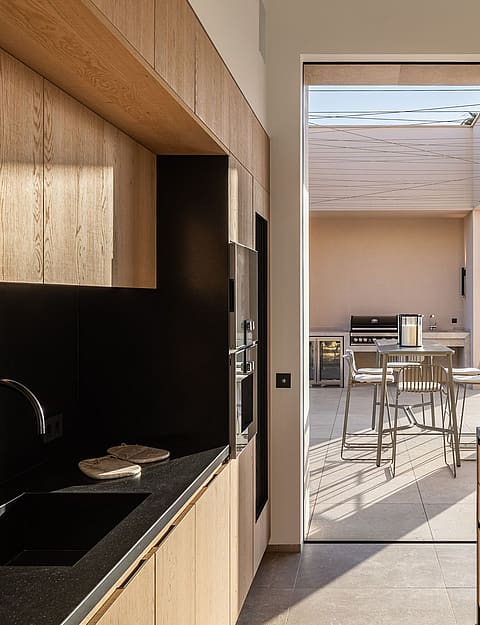 Modern kitchen with natural wood cabinetry and black stone countertops leading through a large opening to an outdoor patio.