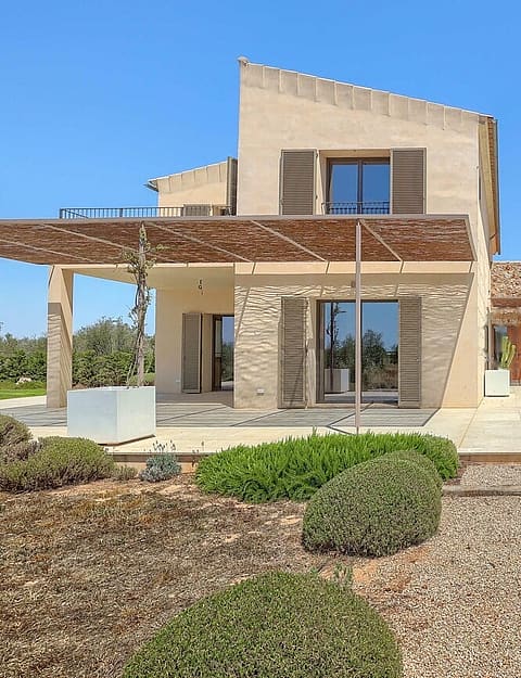 Exterior view of a Mediterranean stone finca featuring a wooden pergola, gravel path, and landscaped garden under a clear sky.