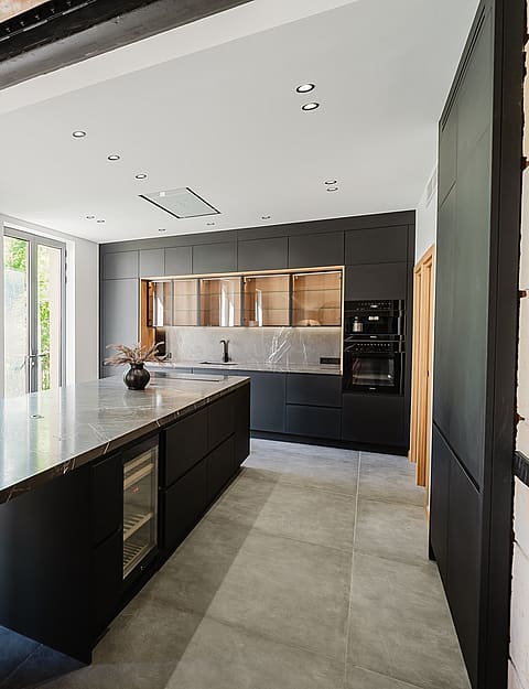 Modern open-plan kitchen in an apartment featuring a large dark stone island with waterfall edges, matte black cabinetry, and exposed wooden ceiling beams.