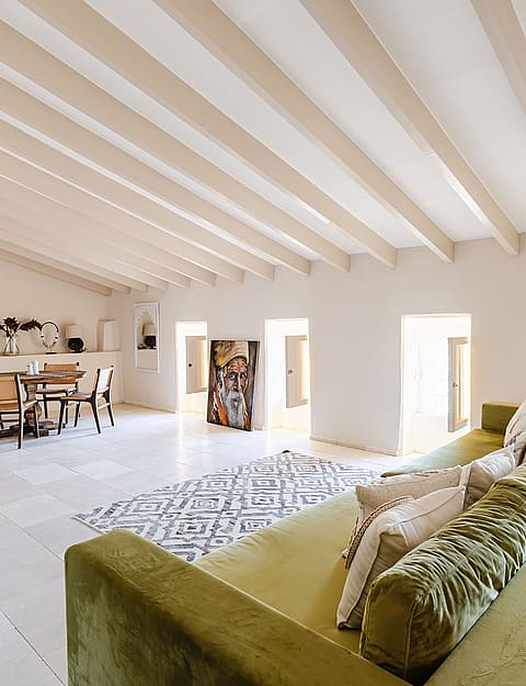 Spacious living room featuring a long olive green sofa, white stone flooring, and exposed wooden beams on a slanted ceiling.