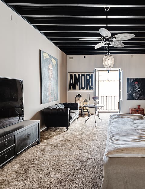 A spacious master bedroom with a white bed, black exposed wood beams, and a small seating area leading to a balcony.