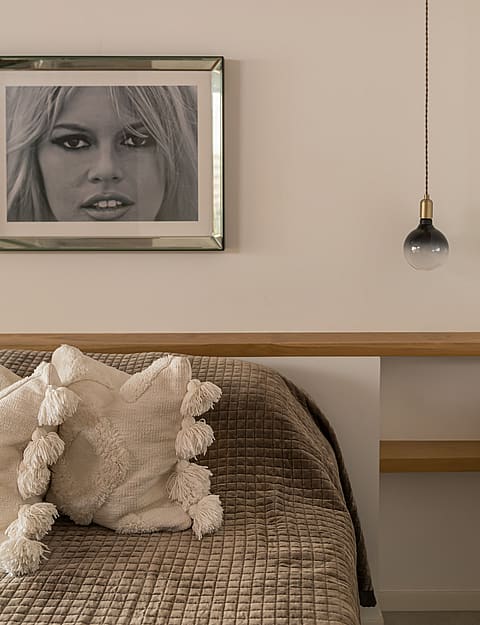 A close-up of a bedroom interior showing a textured brown bedspread, off-white decorative pillows with tassels, and a floating light-oak headboard shelf with a pendant light.