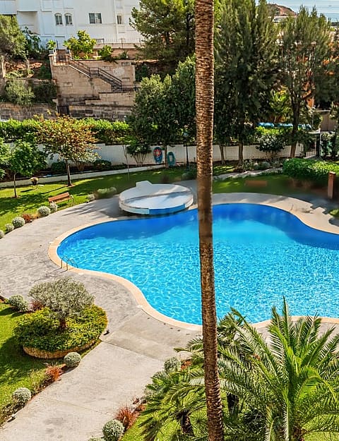 High-angle view of a kidney-shaped blue swimming pool surrounded by manicured green lawns and palm trees in Portals Nous.