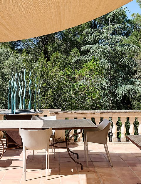 A stone terrace with a dining table and chairs, protected by a large beige sail shade, overlooking dense mountain trees.