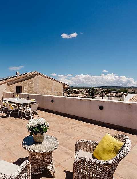 A wide rooftop terrace with stone flooring, wicker outdoor furniture, and a dining table, overlooking the village buildings.