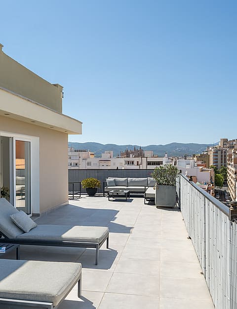 A wide rooftop terrace with light stone flooring, two grey sun loungers in the foreground, and a city skyline view.