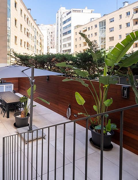 A private outdoor terrace in Palma featuring light-colored stone flooring, wooden wall panels, a dining table with chairs, and an umbrella, set against a backdrop of city buildings.