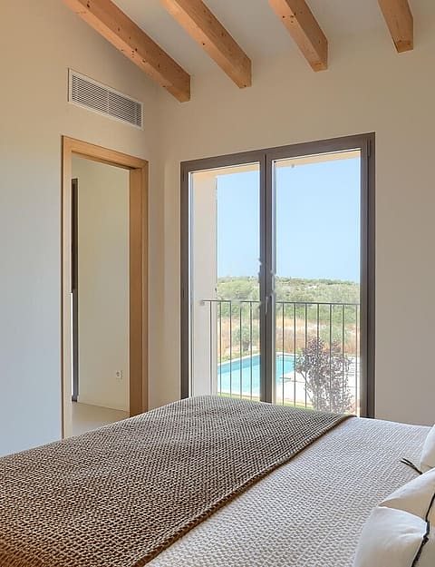 A bedroom featuring exposed timber ceiling beams, white walls, and a glass door opening to a balcony overlooking a pool.
