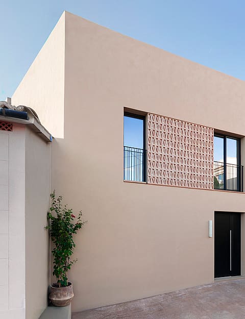 Minimalist sand-colored townhouse facade with a perforated terracotta screen, dark entrance door, and integrated planters.