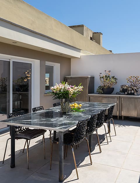 A spacious outdoor terrace with a long dining table, modern chairs, and an open view overlooking the city and the sea.