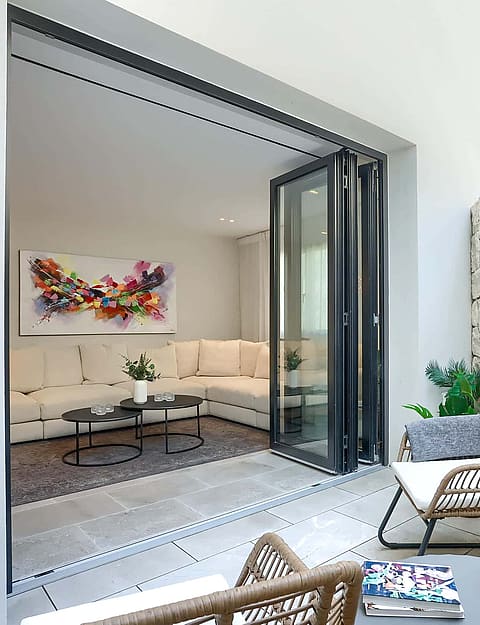 A small outdoor terrace with a high natural stone wall, rattan chairs, and large folding glass doors opening into a living room.