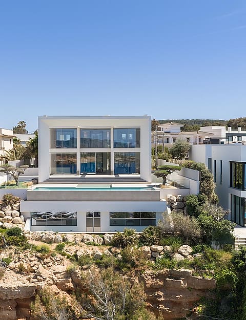 Wide exterior view of a modern white multi-level villa built into a rocky coastline with a swimming pool and glass walls.