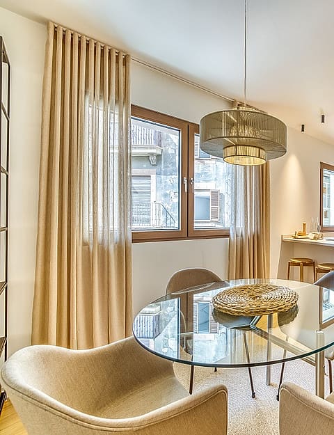 Modern open-plan kitchen with white cabinetry and dining area featuring a round glass table, beige armchairs, and floor-length curtains.