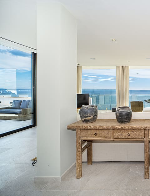 Interior view of an open-plan apartment featuring a rustic wooden console table, white walls, and floor-to-ceiling glass doors opening to a sea-facing terrace.