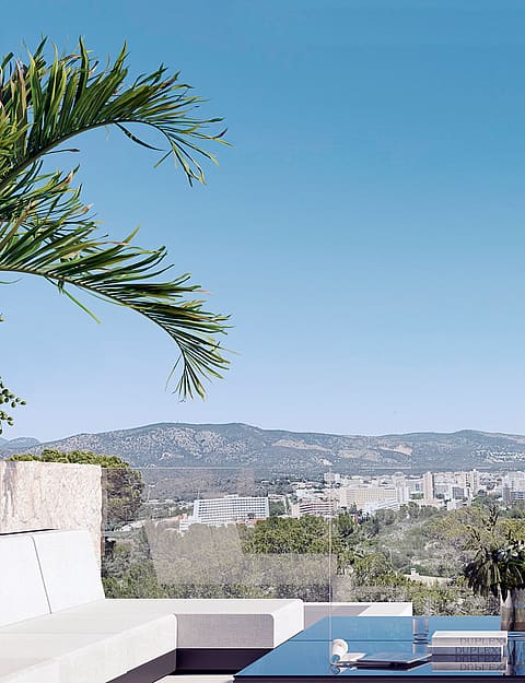 An outdoor terrace with white minimalist sofa and glass-topped table overlooking a valley, cityscape, and mountain range.