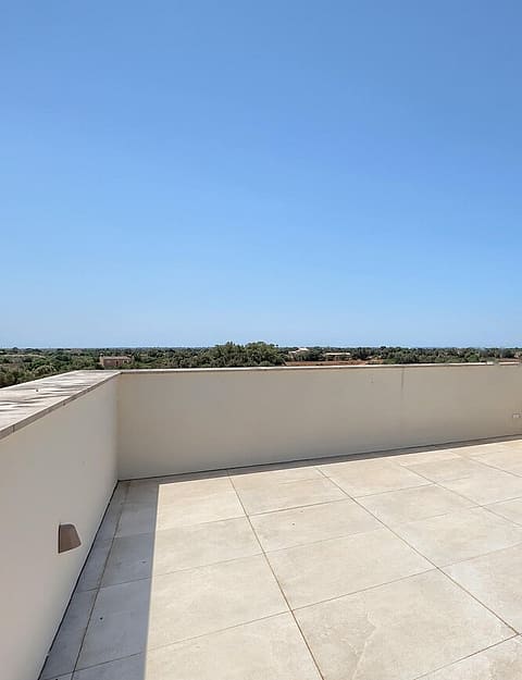 Empty rooftop terrace with pale stone tiling and low plastered walls under a clear blue sky, overlooking distant countryside.