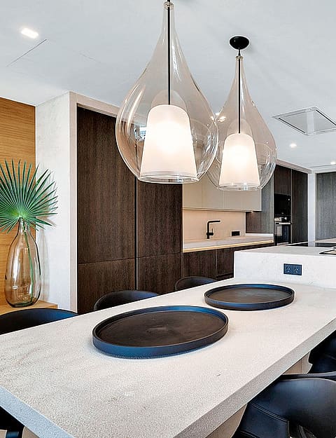 Modern open-plan kitchen featuring a long light-colored stone island, dark cabinetry, and two clear glass pendant lights.
