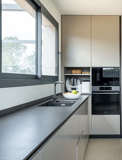 Modern kitchen featuring matte grey cabinetry, dark stone countertops, integrated stainless steel appliances, and large window.