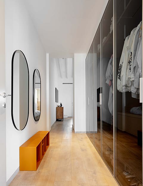A bright hallway featuring light wood flooring, a long wall of dark glass-fronted wardrobes, and minimalist wall mirrors.