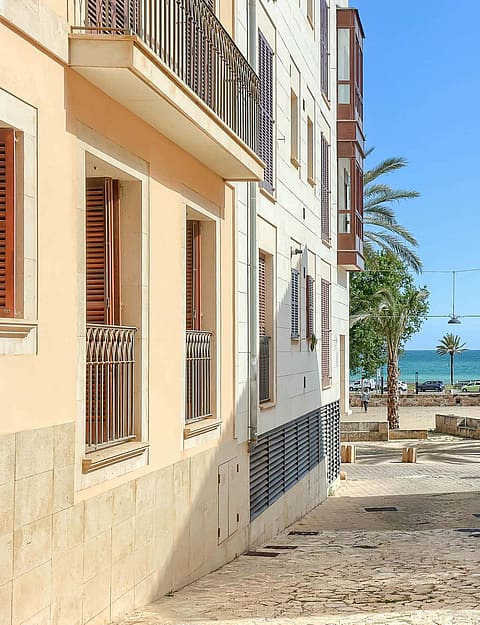 A narrow cobblestone street in a historic Old Town, framed by pale-colored residential buildings with wooden shutters, leading directly to a blue sea view.