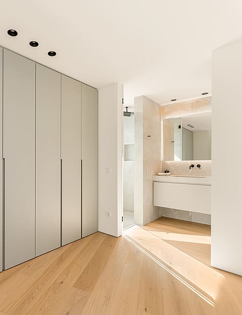 A modern bedroom featuring a floor-to-ceiling grey wardrobe wall, light oak wood flooring, and a view into a bright bathroom.