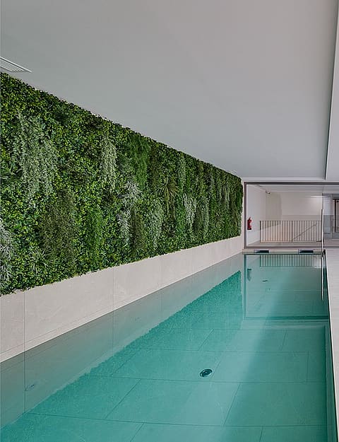 Piscina interior con un largo jardín vertical en una de las paredes y pavimento minimalista gris.