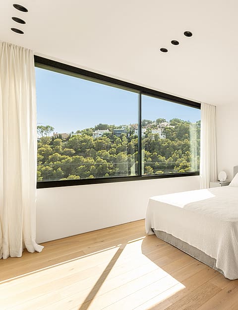Modern bedroom with oak wood flooring, grey upholstered bed, and floor-to-ceiling windows showing hillside pine trees.